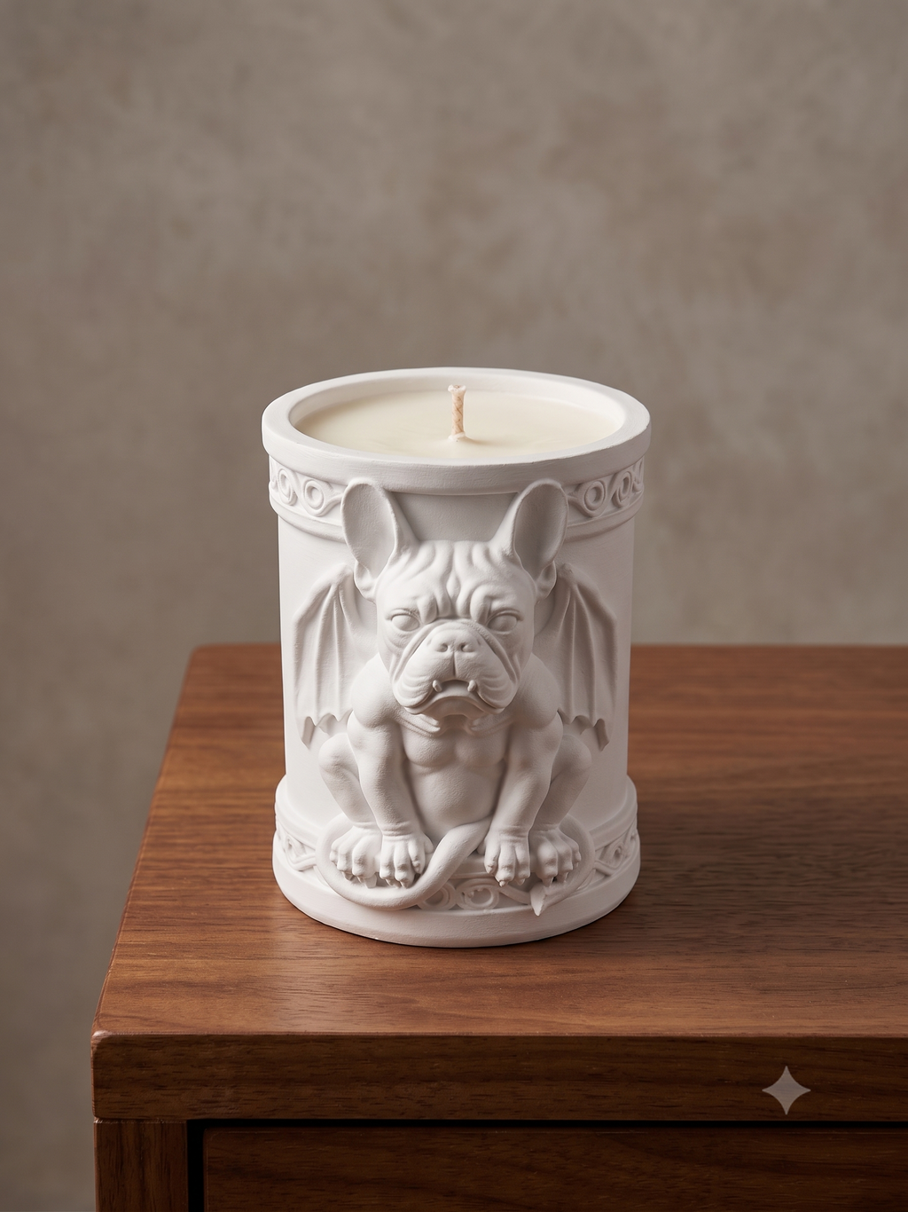 A white ceramic candle jar featuring a sculpted bulldog with bat wings, displayed on a wooden surface against a textured beige wall.