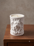 A white ceramic candle jar featuring a sculpted bulldog with bat wings, displayed on a wooden surface against a textured beige wall.